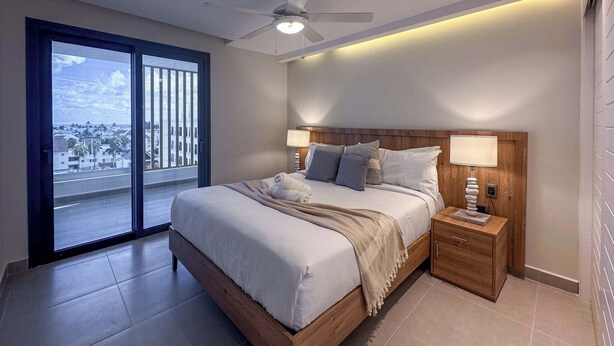 Un dormitorio moderno con una cama grande, dos mesitas de noche, un ventilador de techo y vistas a través de un ventanal corredero.