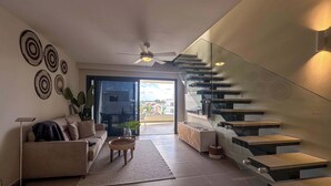 Una sala de estar moderna con sofá, mesa de centro y decoración en la pared. Una escalera con barandilla de cristal conduce a un balcón con vista a la ciudad.