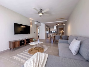 A modern living room with a grey sofa, a flat-screen TV, a wooden entertainment unit, and a dining area with a table and chairs.