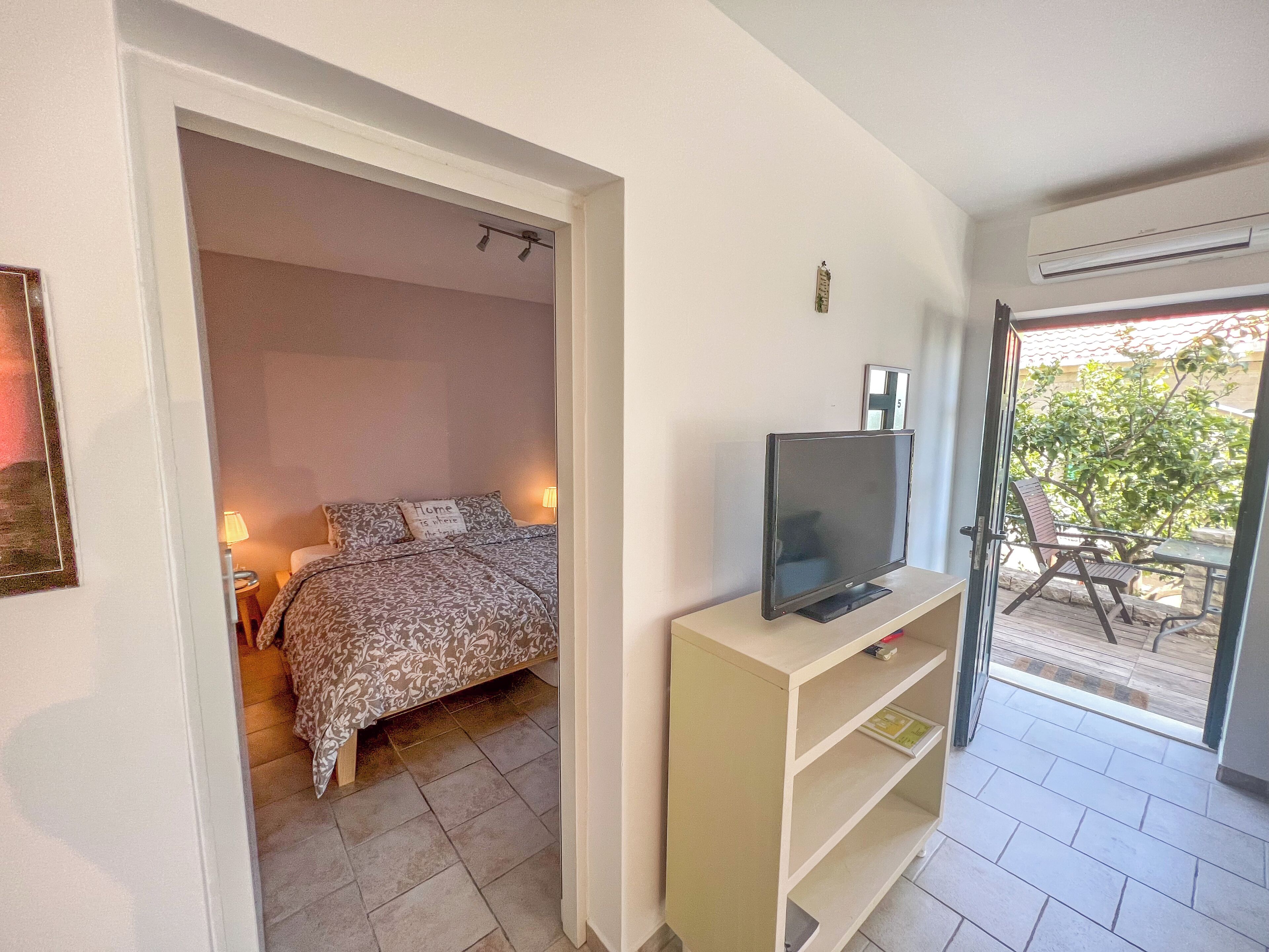 Et soveværelse med seng, et fjernsyn på et stativ og en dør, der fører til en balkon med havemøbler.