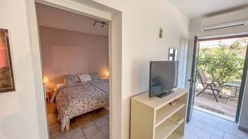 Une chambre à coucher avec un lit, un téléviseur posé sur un meuble et une porte donnant sur un balcon aménagé avec du mobilier d’extérieur.