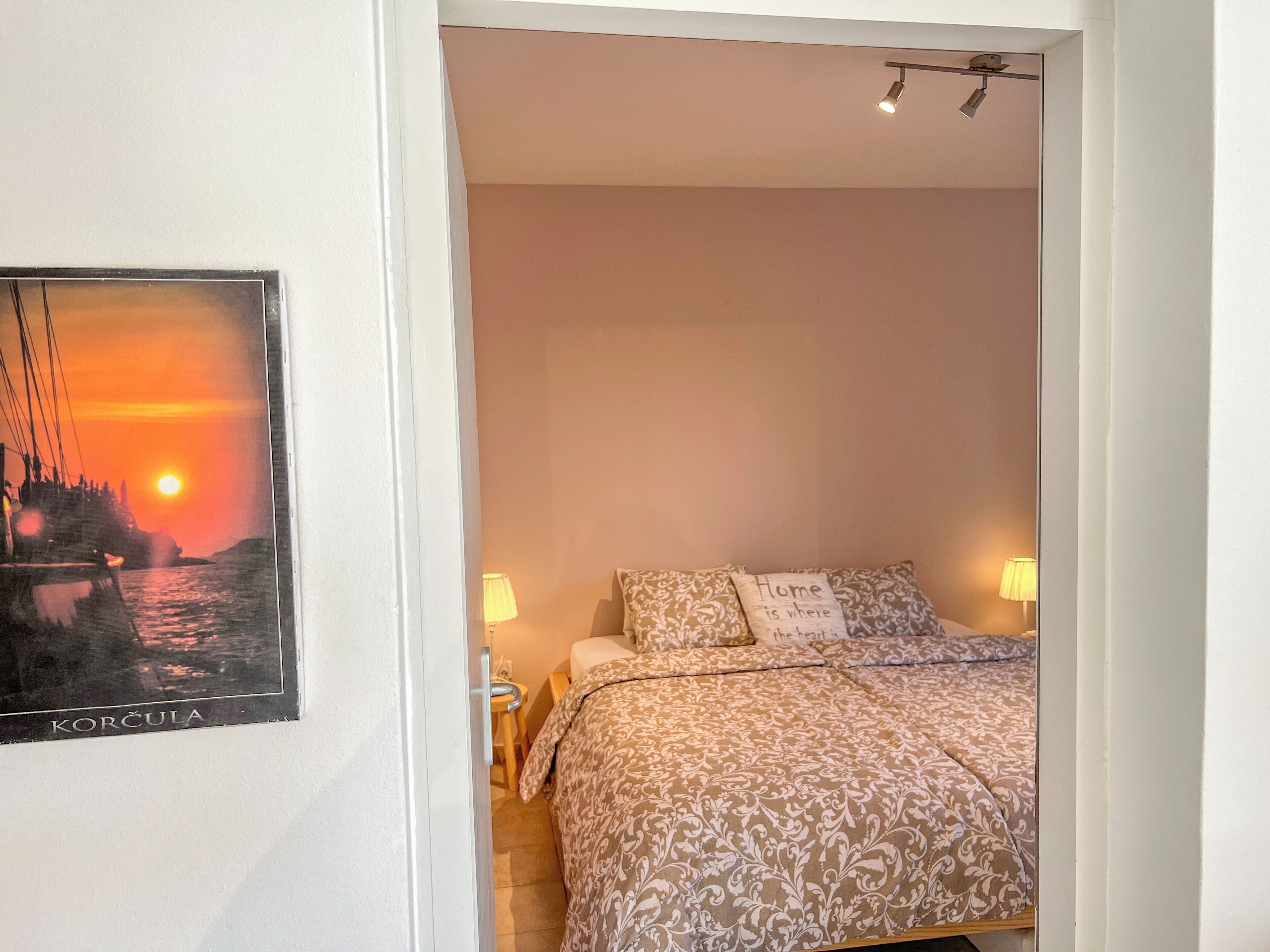 A bedroom with a bed, two bedside lamps, a pillow with a quote, and a framed picture on the wall.