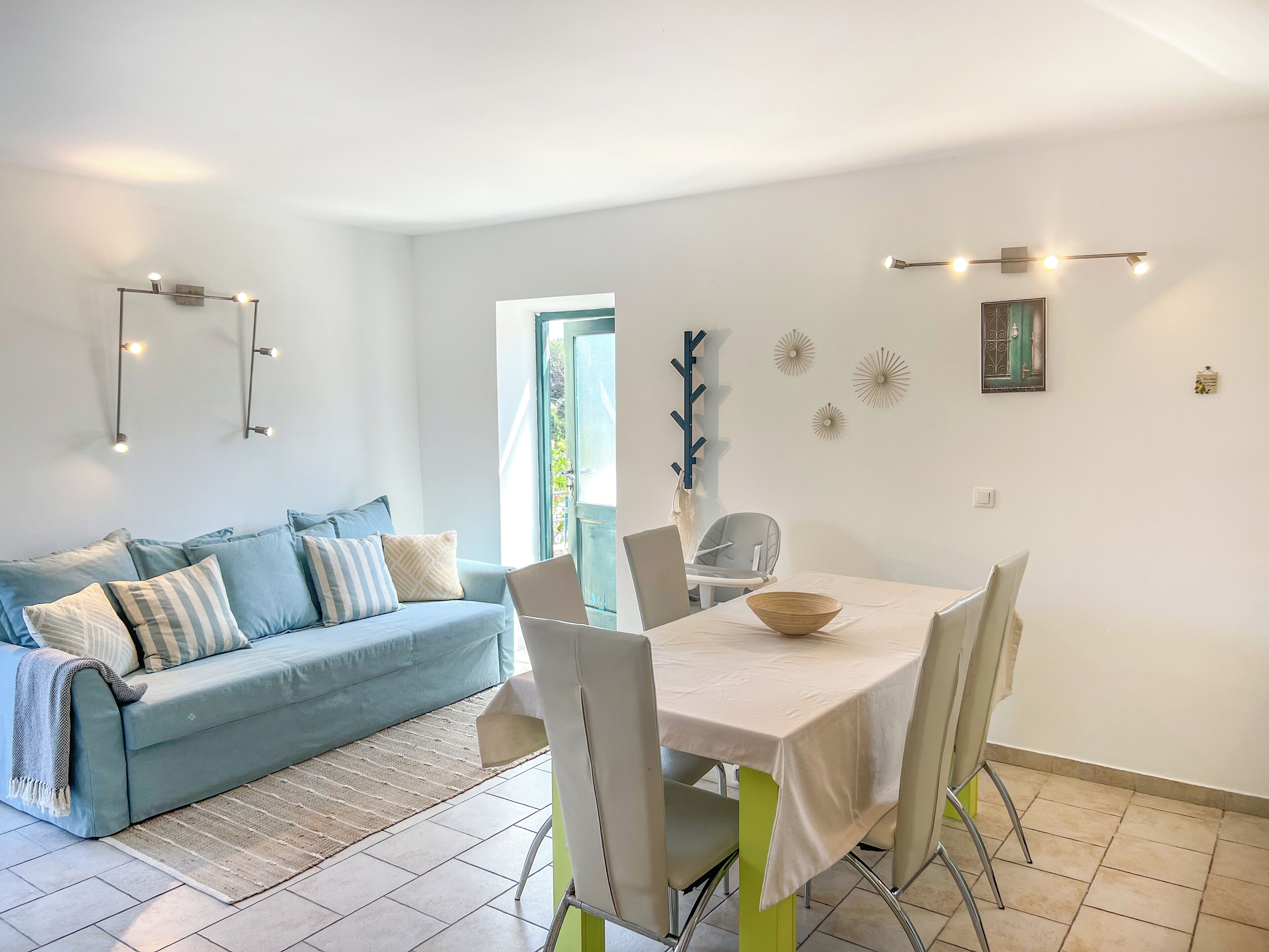 A modern living room with a light blue sofa, a dining table with chairs, and a window with a view of greenery.
