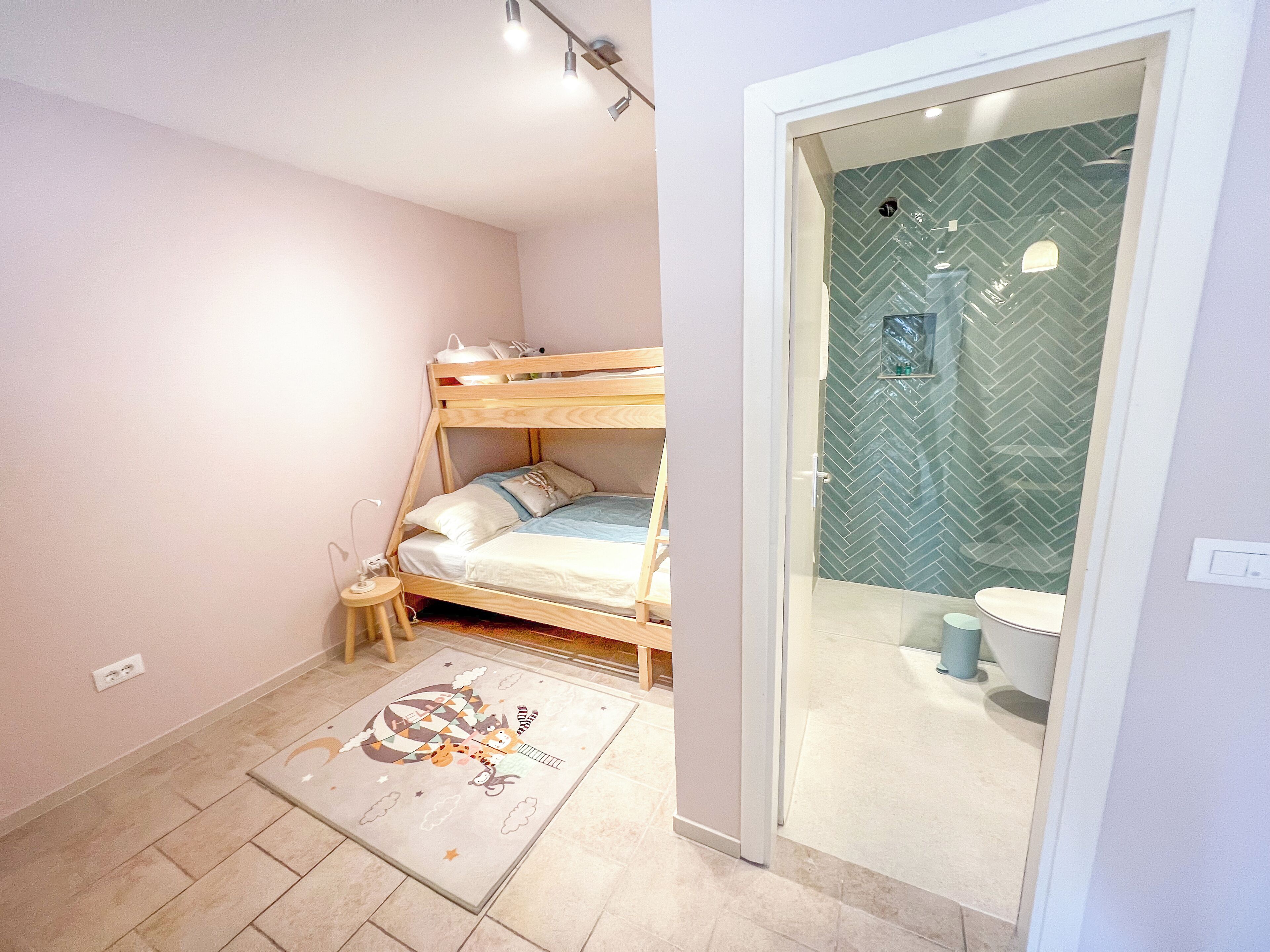 Un dormitorio con una cama litera, una mesita de noche pequeña, un tapete y un baño con un salpicadero de azulejos tipo espiga.