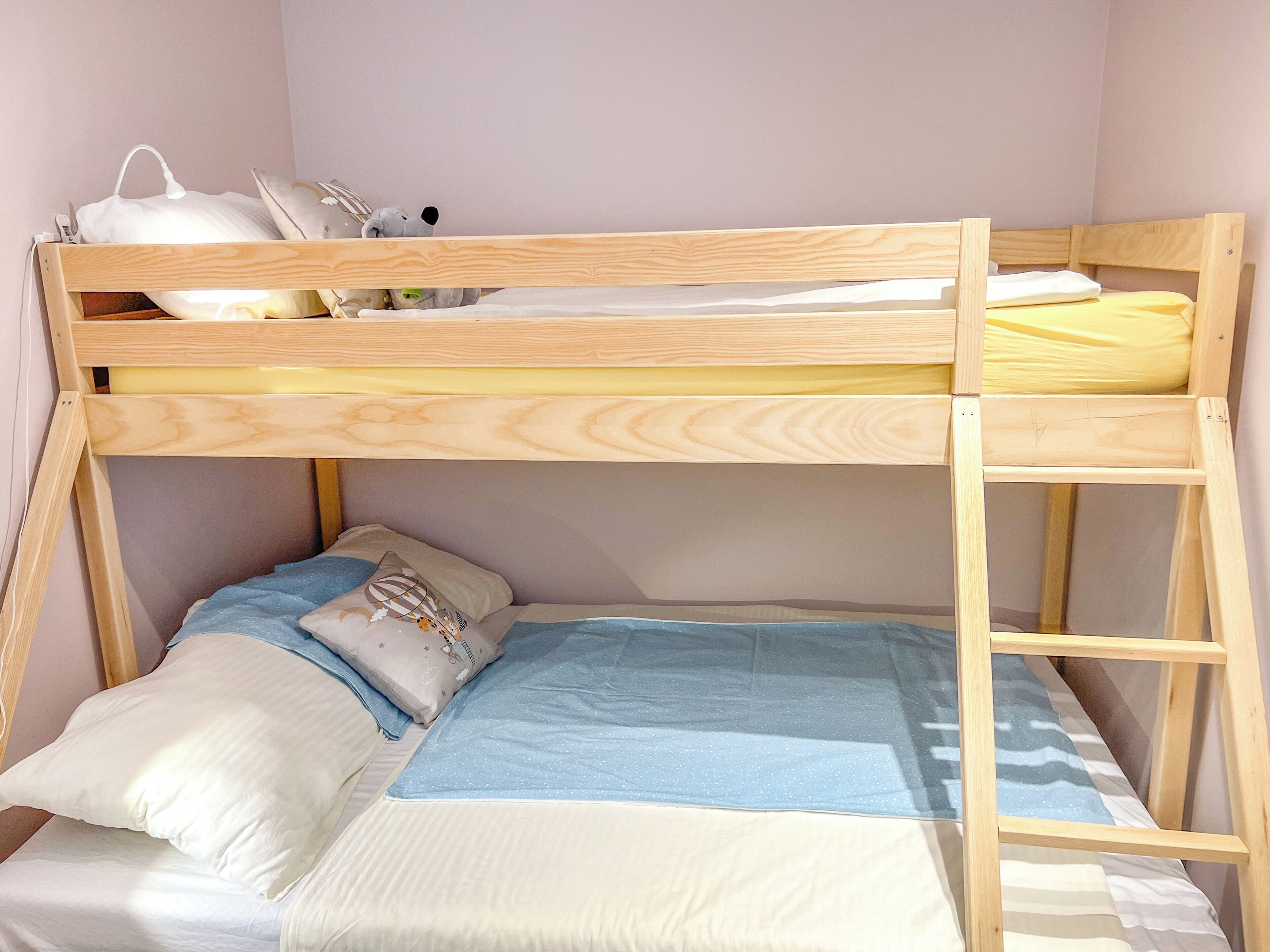 Ein Stockbett mit einem unteren und einem oberen Bett. Das untere Bett hat eine blau-weiße Matratze, das obere eine gelbe. Auf dem oberen Bett liegen Kissen und ein Stofftier.