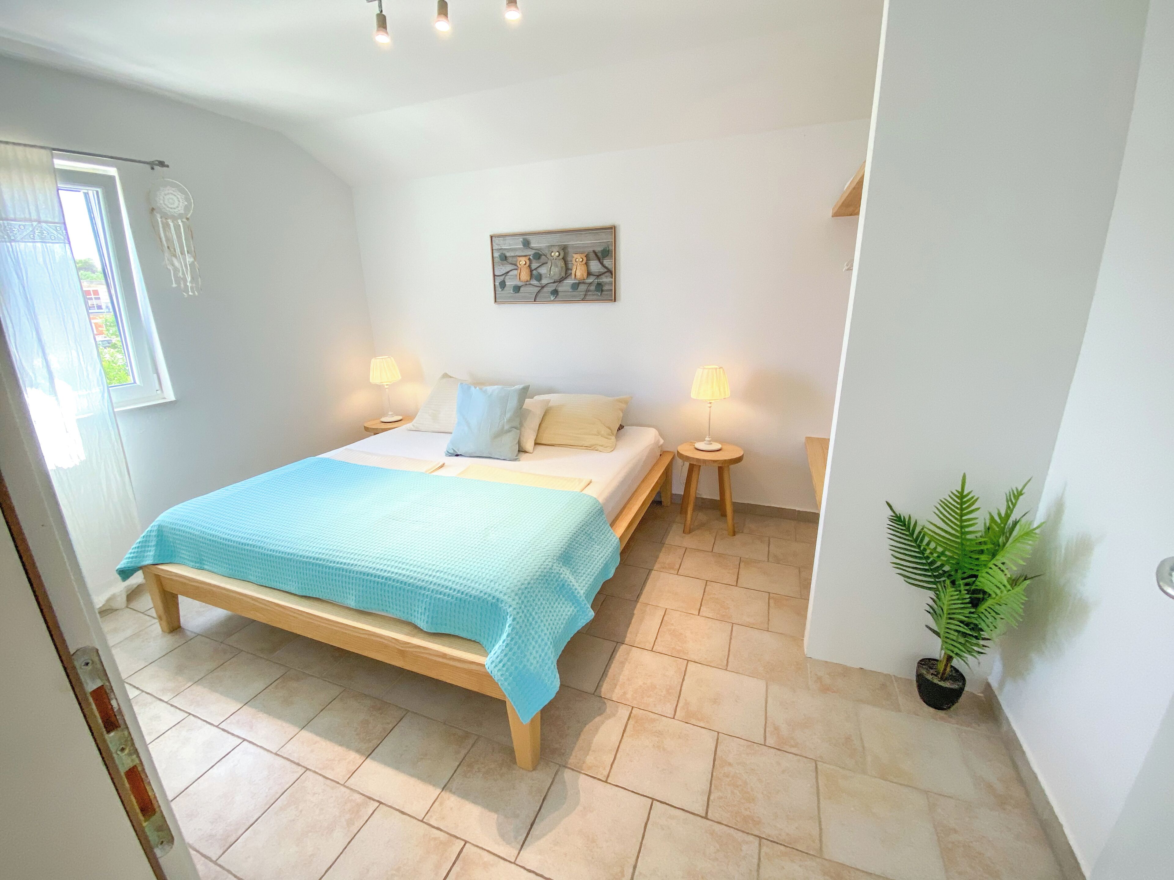 A bedroom with a bed, bedside lamps, a small table, a potted plant, and a wall-mounted artwork.