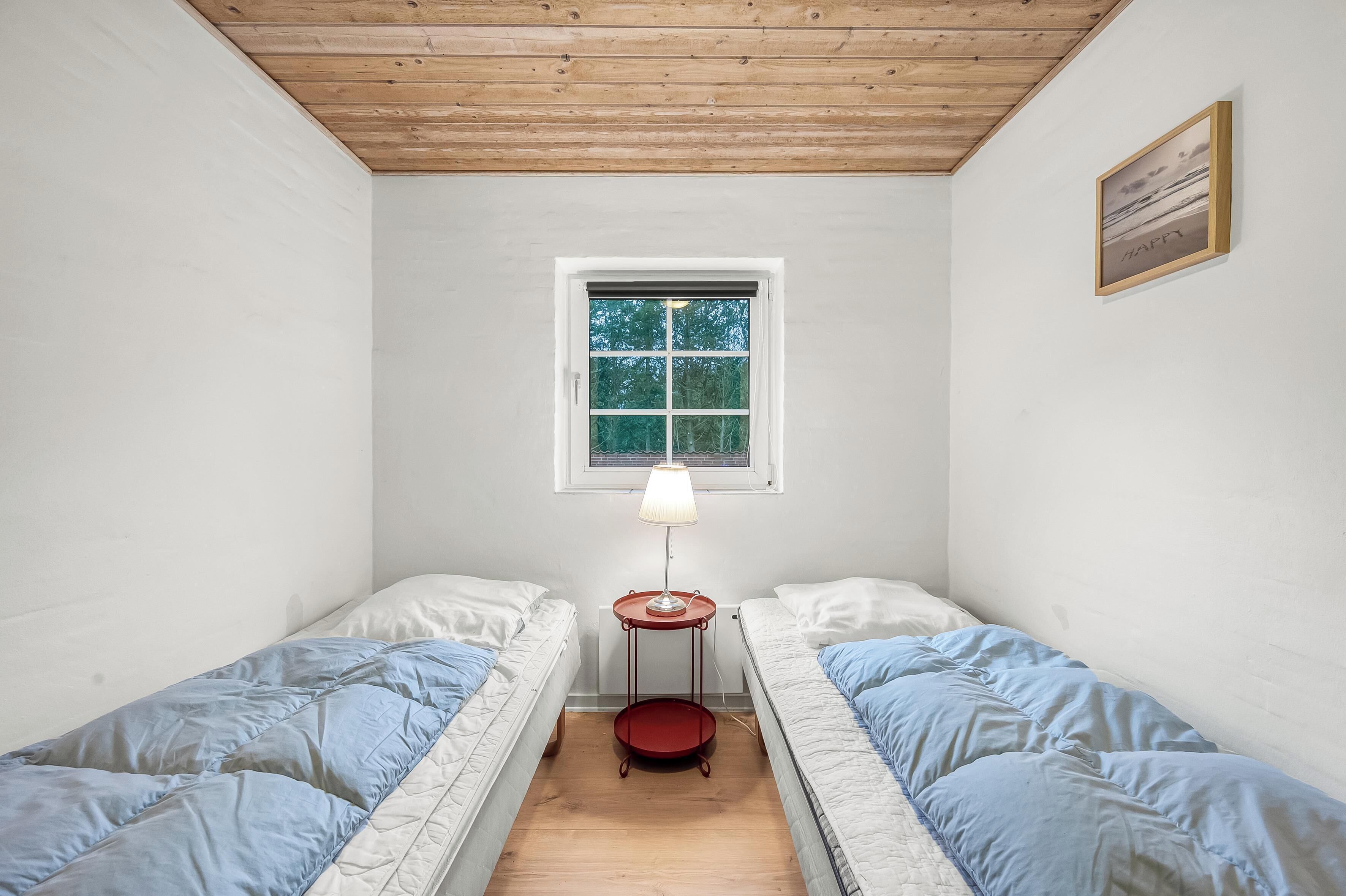A small, well-lit room with two single beds, a small table with a lamp, and a window providing natural light.