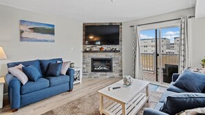 Un salon avec un canapé bleu, une cheminée, un téléviseur fixé sur un mur en pierre et une vue sur l’extérieur par une porte vitrée.