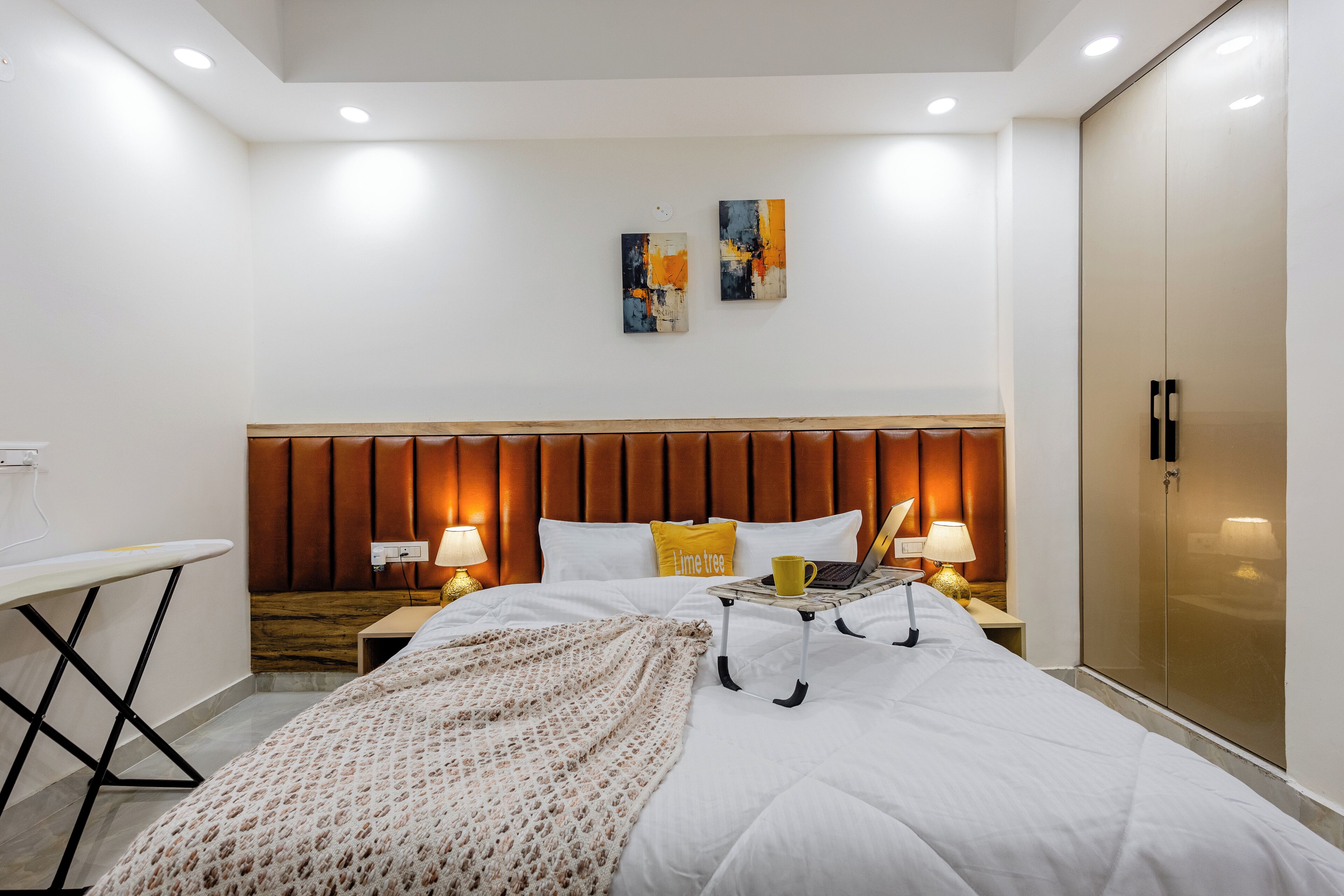 Een moderne hotelkamer met een houten hoofdeinde, een bed met witte lakens, een nachtkastje met een lamp, een laptop en een koffiekop. Twee abstracte schilderijen hangen aan de muur en er staat een kledingkast met spiegel in de kamer.