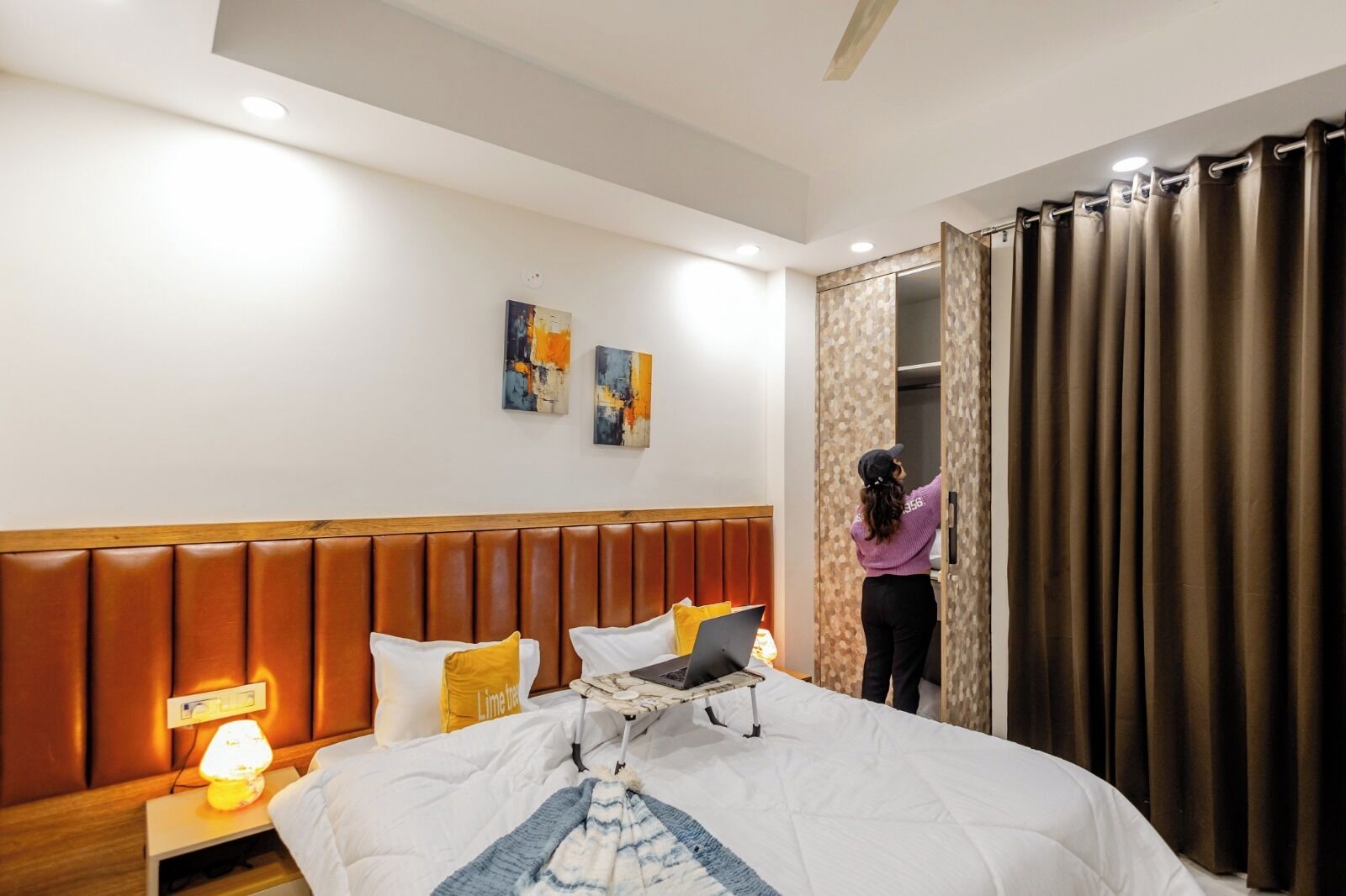 Una habitación de hotel con cama, mesita de noche, lámpara, una obra de arte en la pared y una persona ajustando las cortinas.