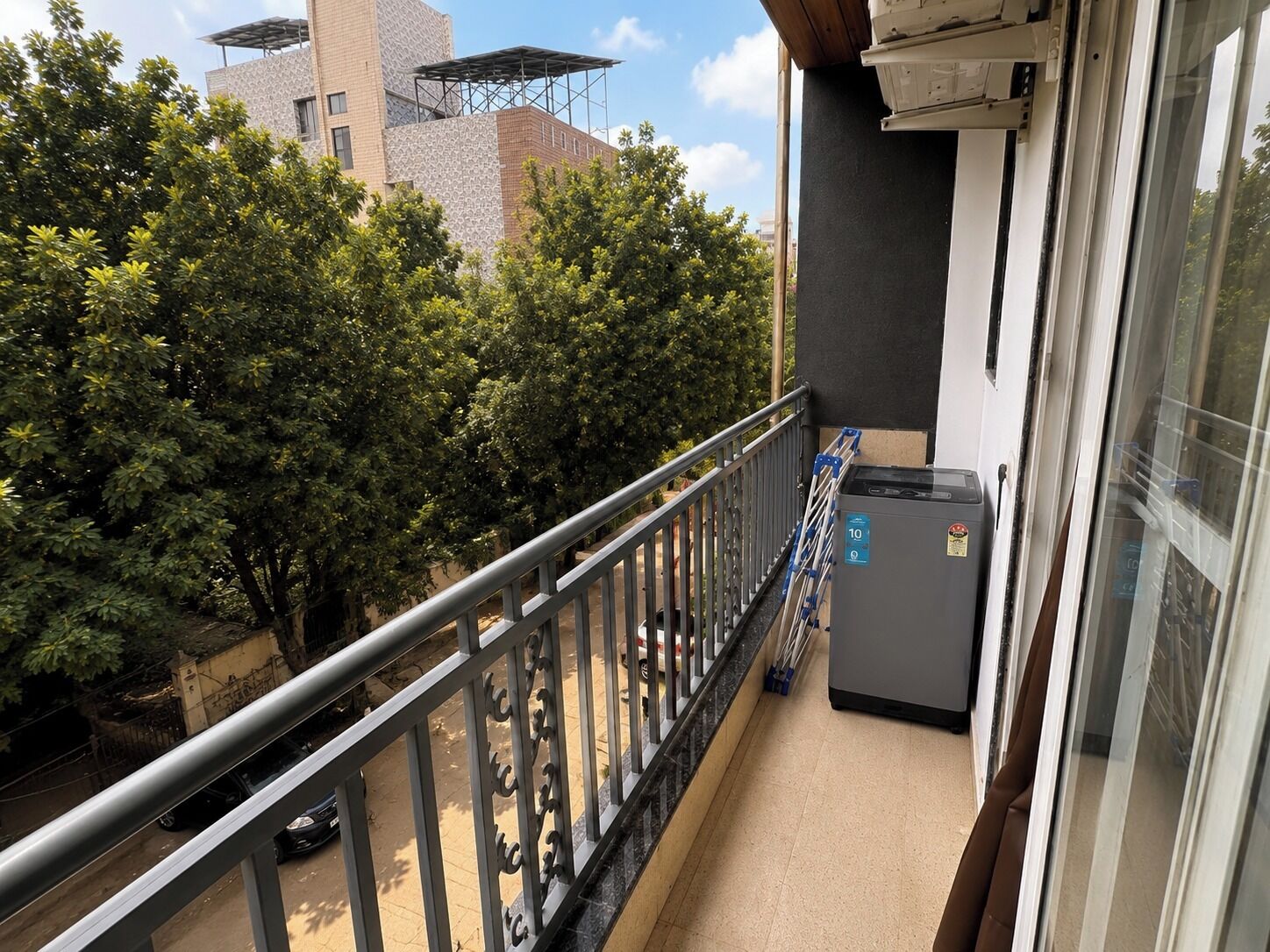 Een balkon met een metalen hekwerk, een grijze airconditioning en uitzicht op bomen en gebouwen.