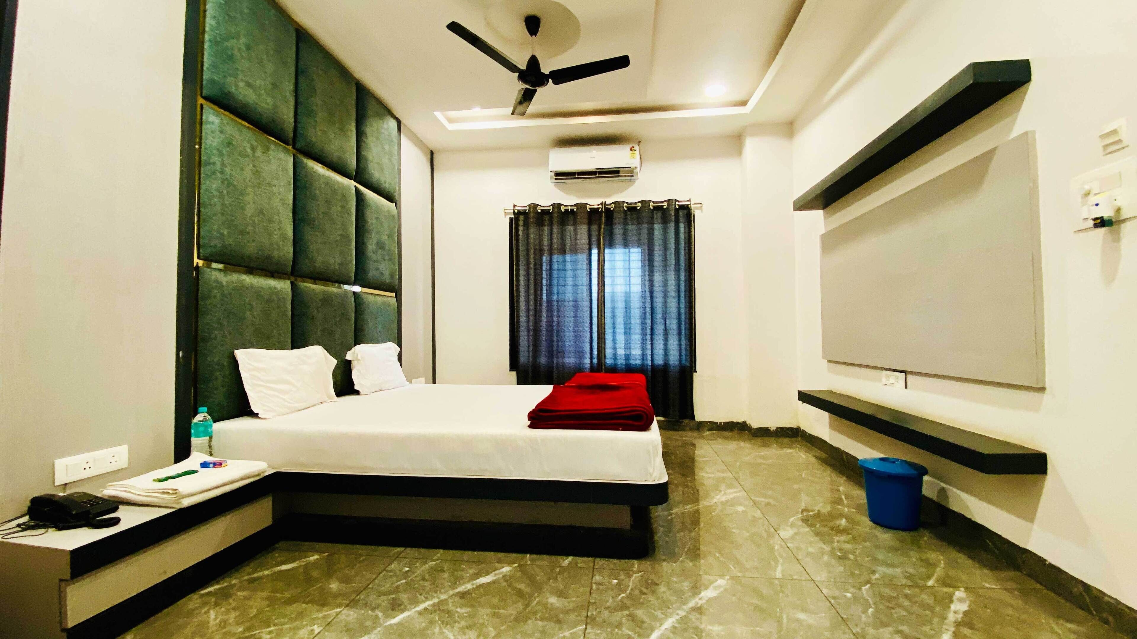 A hotel room with a bed, a desk, a phone, a water bottle, a ceiling fan, an air conditioner, a TV, and a window with curtains.