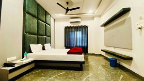 A hotel room with a bed, a desk, a phone, a water bottle, a ceiling fan, an air conditioner, a TV, and a window with curtains. - Hotel JK Palace (Khandwa)