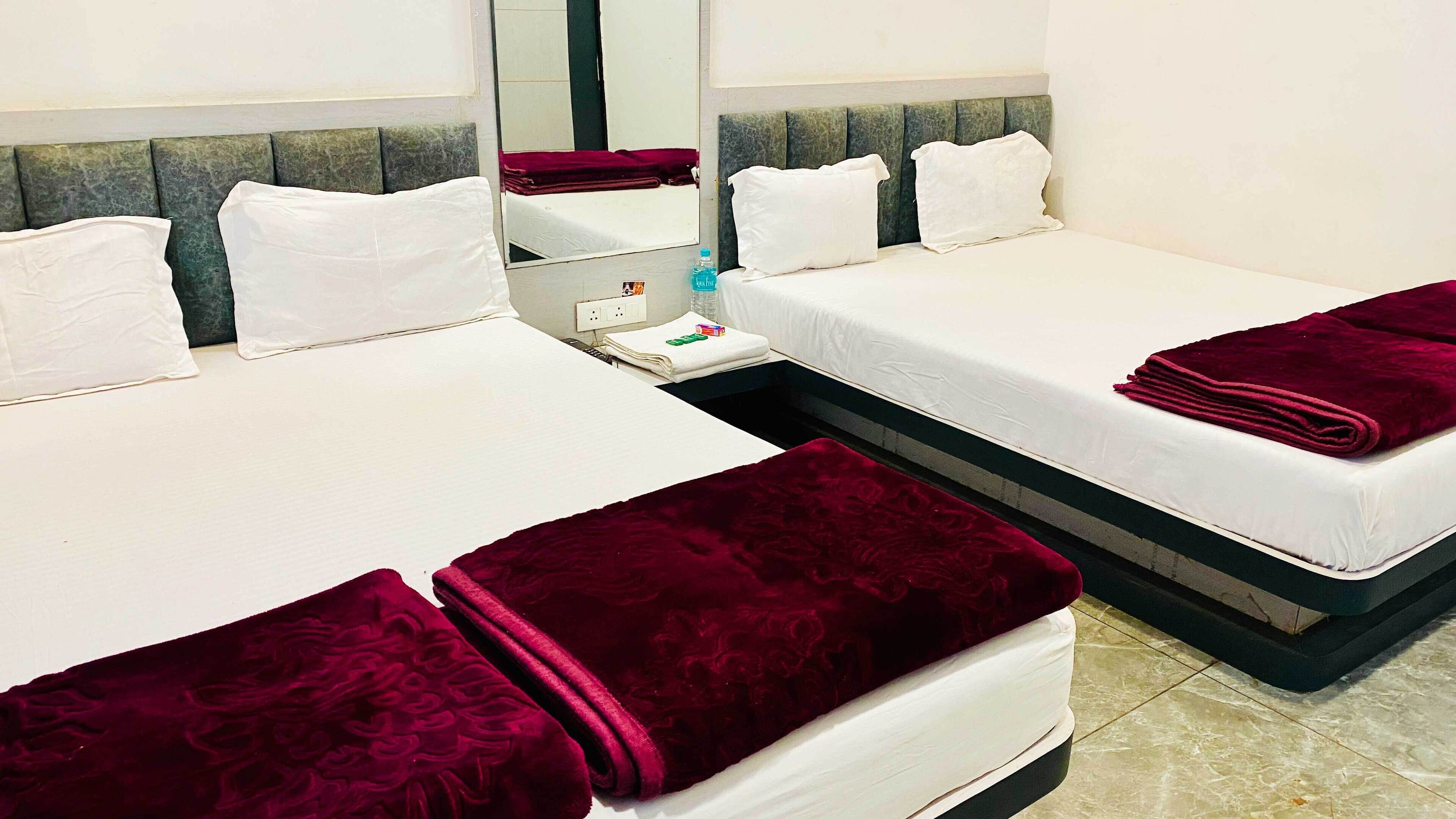 A hotel room with two beds, each with white linens and a dark red blanket. There is a nightstand with a water bottle and a small item on it. The room has a modern design with a grey headboard and a large mirror above the beds.
