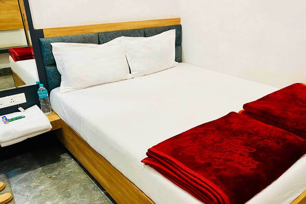 Ein ordentlich bezogenes Bett mit weißen Leinen und einer roten Decke, ein hölzerner Kopfseitenrahmen und ein Nachttisch mit einer Wasserflasche und einer Taschentuchbox.