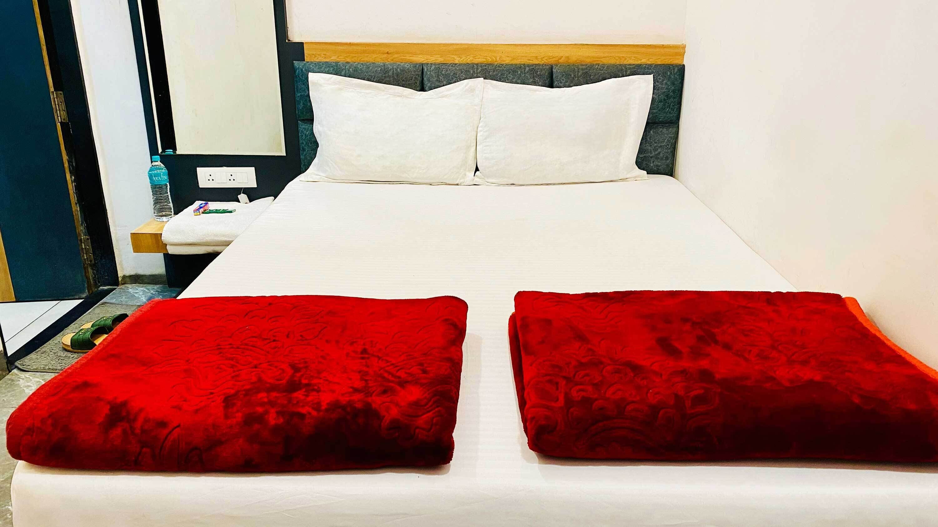 Ein Hotelzimmer mit Bett, zwei Kissen, einem Nachttisch, einer Wasserflasche, Hausschuhen und einem kleinen Tisch.