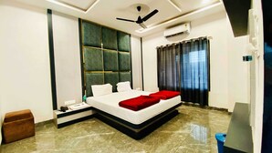 A hotel room with a bed, a ceiling fan, an air conditioning unit, a brown ottoman, and a blue trash bin. - Hotel JK Palace (Khandwa)