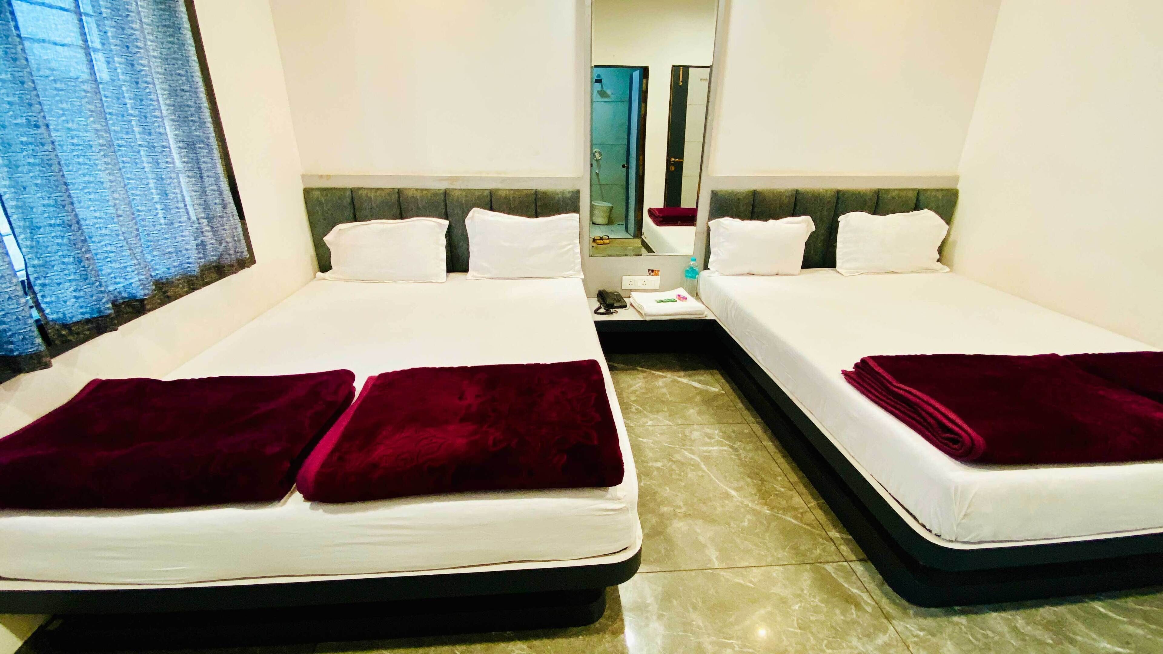 A hotel room with two beds, each with white linens and a dark red throw blanket. There is a small table with a phone and a bottle of water. The room has a tiled floor and a window with curtains.