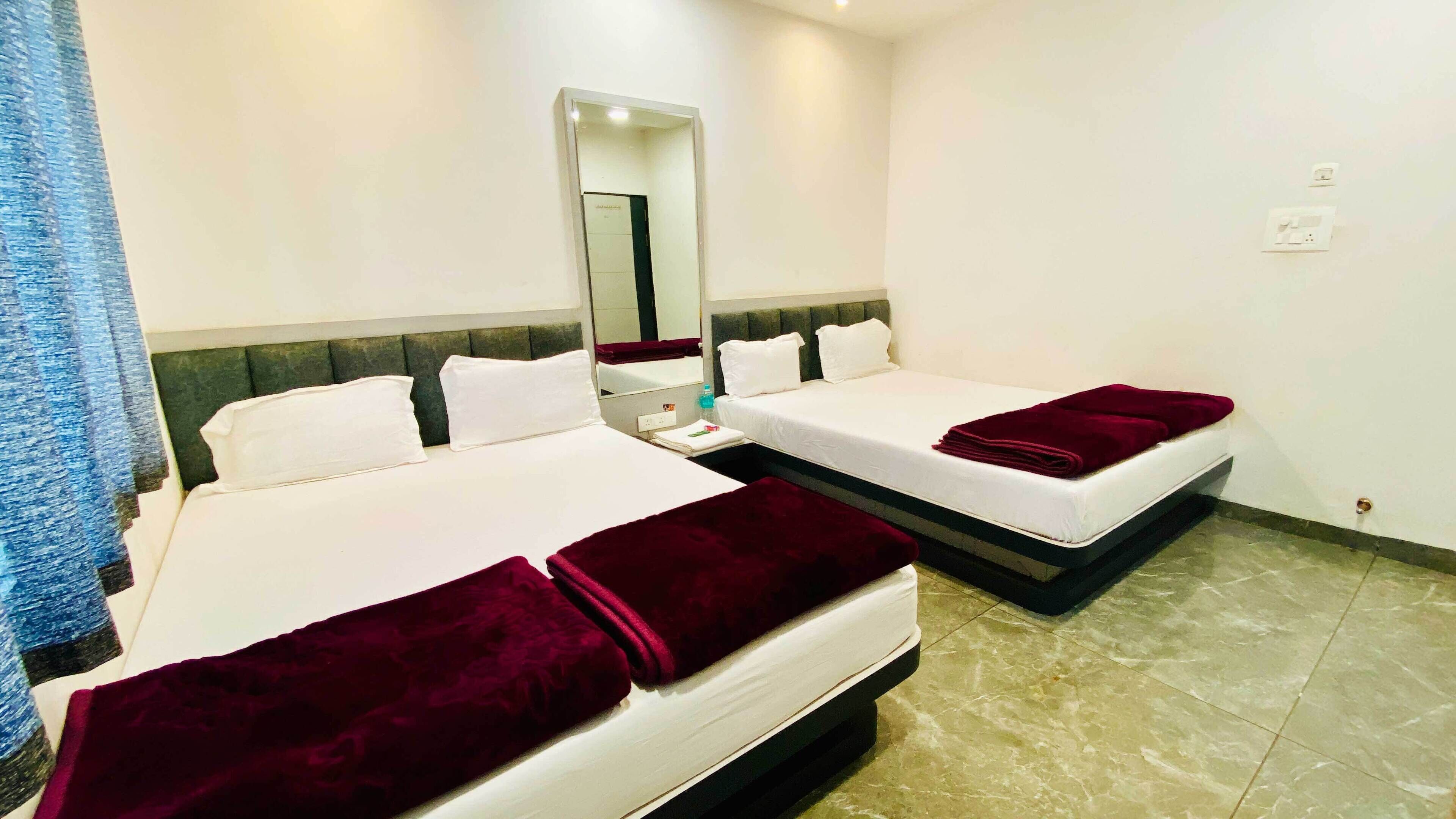 A hotel room with two beds, each with white linens and a dark red blanket. There is a mirror above the beds, a small table with a bottle and a tissue box, and a light switch on the wall.