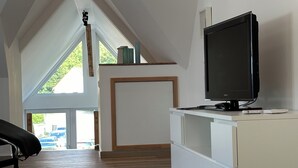 Ein geräumiger Raum mit schrägem Dach, einem Fernseher auf einem weißen Schrank und Blick auf eine grüne Landschaft durch ein großes Fenster.