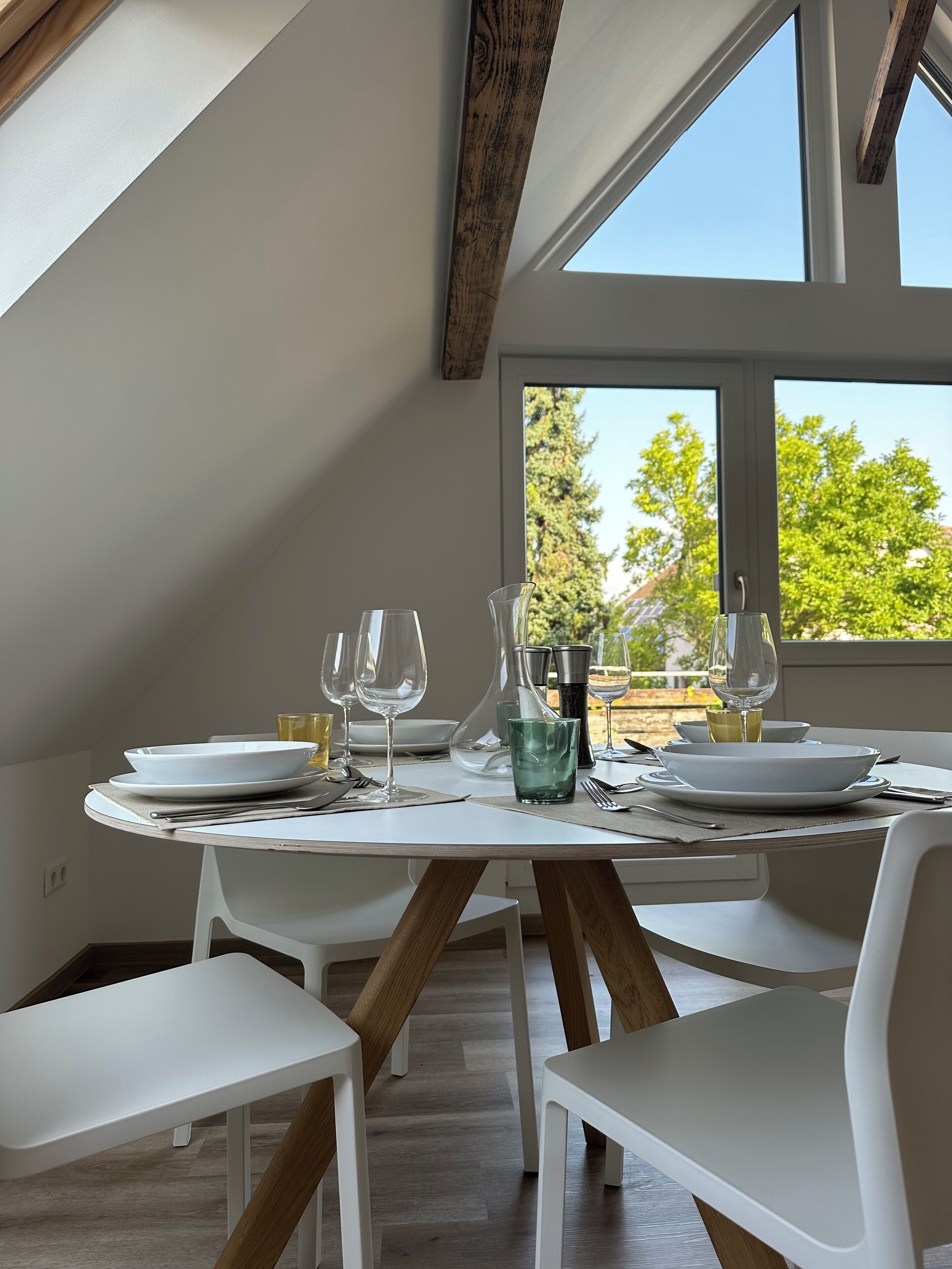 A dining area with a round table set for four, white chairs, and a view of trees through large windows.