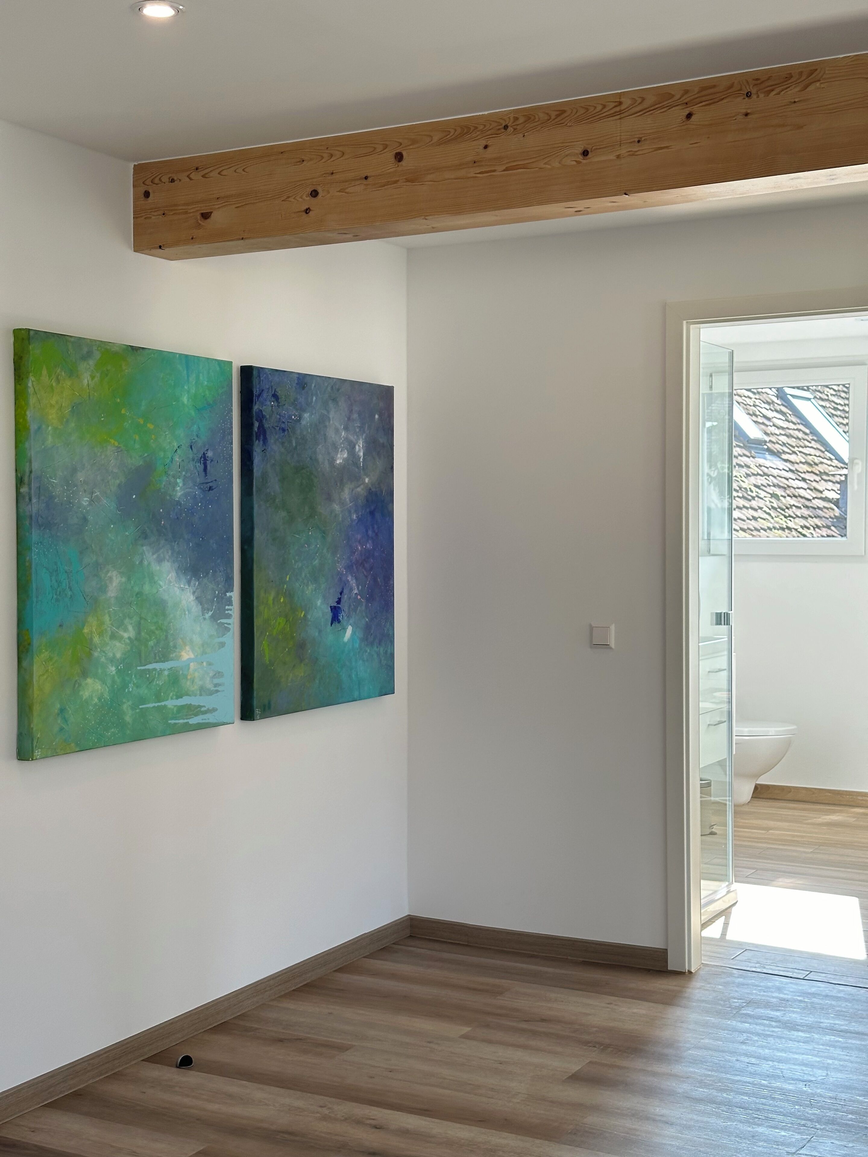 Quarto com piso de madeira, vigamento de madeira no teto e duas pinturas abstratas na parede.