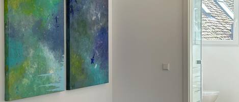 Quarto com piso de madeira, vigamento de madeira no teto e duas pinturas abstratas na parede.