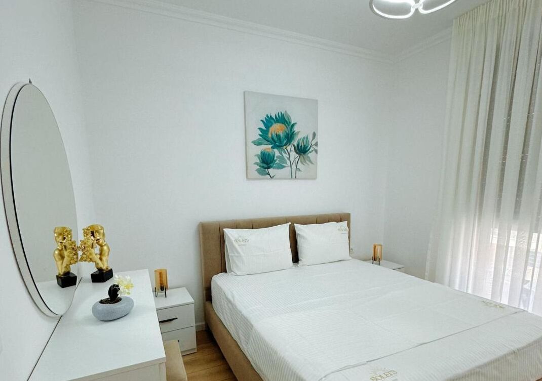 Un dormitorio con cama, mesita de noche, espejo y una obra de arte colgada en la pared.