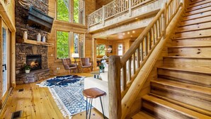 A log cabin interior with a stone fireplace, wooden staircase, and large windows.
