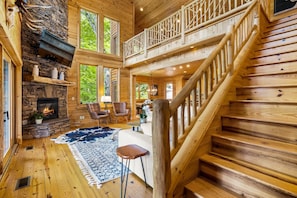 A log cabin interior with a stone fireplace, wooden staircase, and large windows.