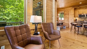 A wooden cabin interior with two leather chairs, a table, a lamp, and a kitchen area with a microwave.