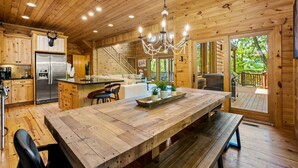 A wooden cabin interior with a large wooden dining table, a chandelier, a refrigerator, and a staircase leading to a deck.