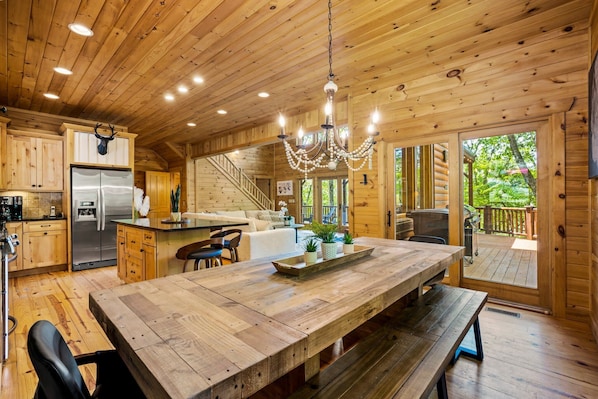A wooden cabin interior with a large wooden dining table, a chandelier, a refrigerator, and a staircase leading to a deck.
