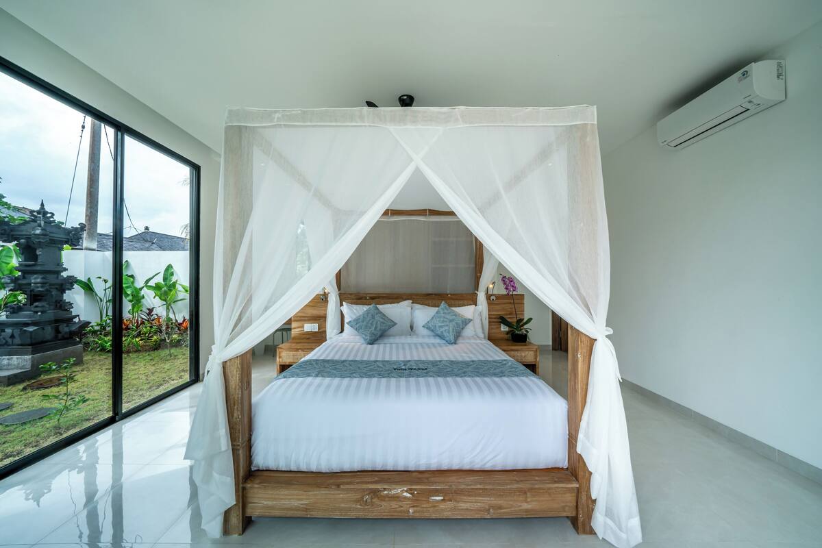 Cama com dossel branco, cabeceira de madeira e duas almofadas.