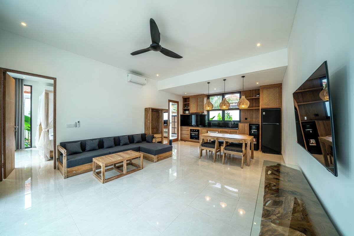 Ampla sala de estar com sofá, mesa de centro e mesa de jantar. Cozinha com armários de madeira e uma grande TV fixada na parede.