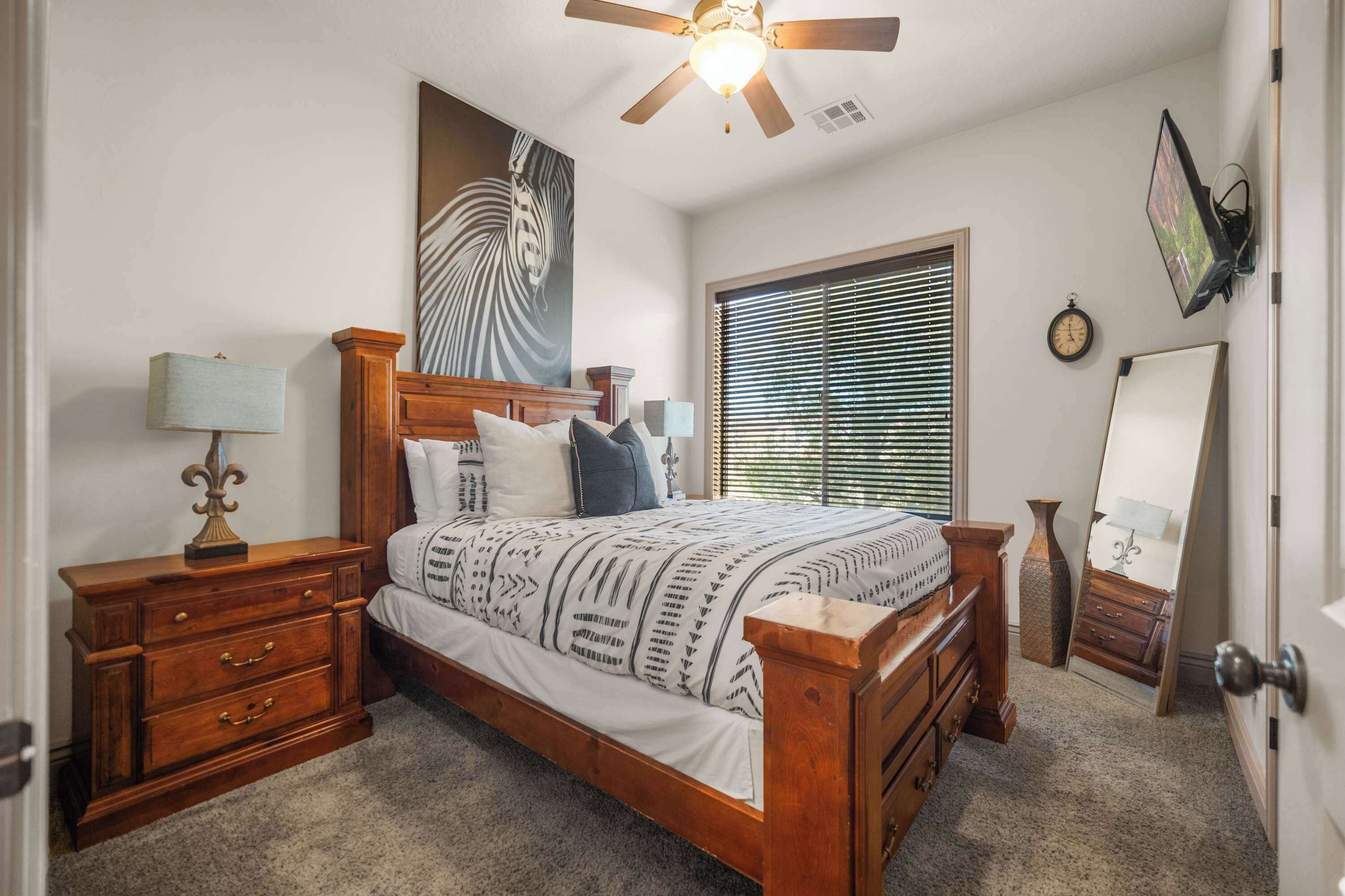 A bedroom with a wooden bed, a nightstand, a dresser, a ceiling fan, a window with blinds, and a wall clock.