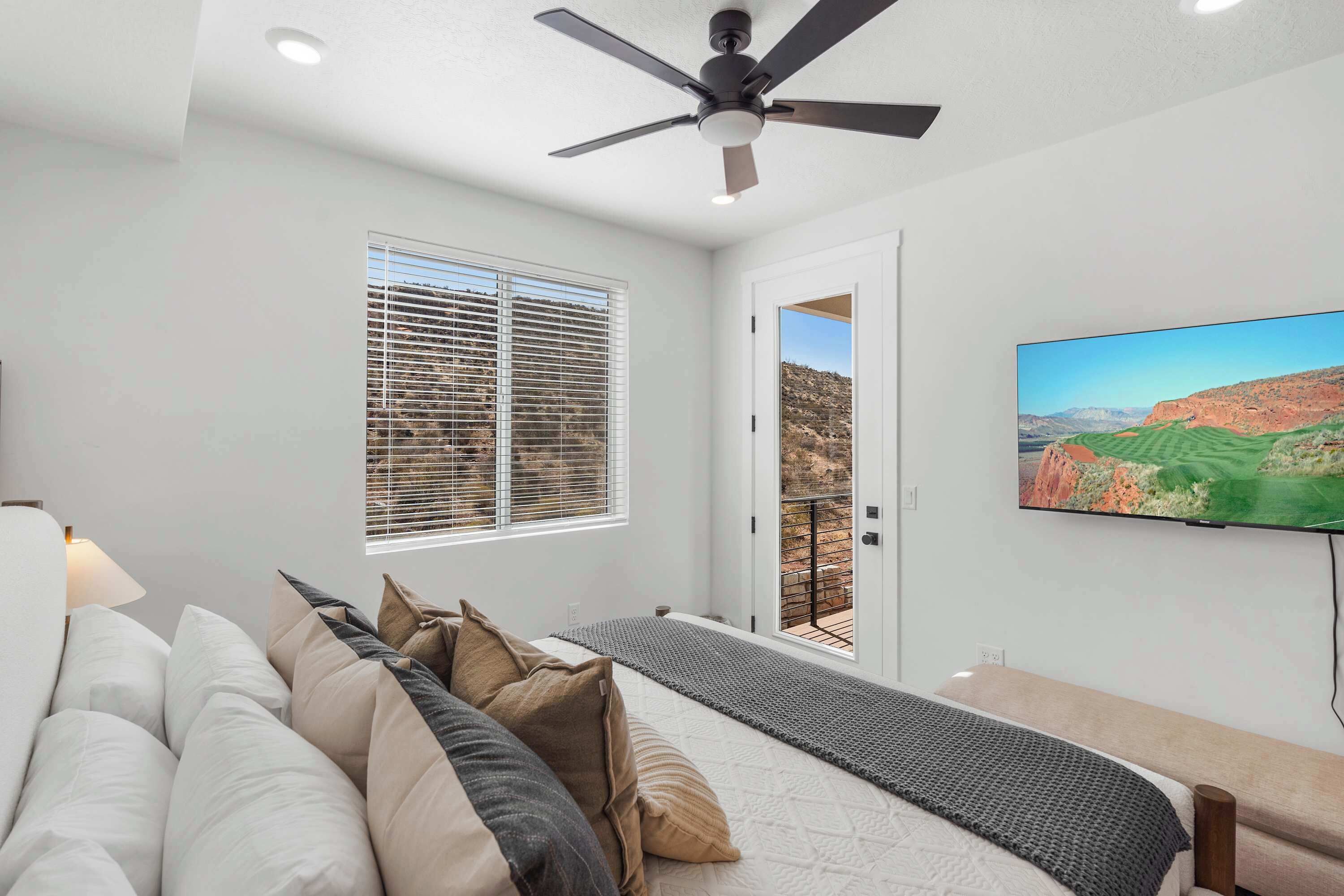 A bedroom with a bed, a ceiling fan, a window with blinds, a door leading to a balcony, and a TV on the wall.