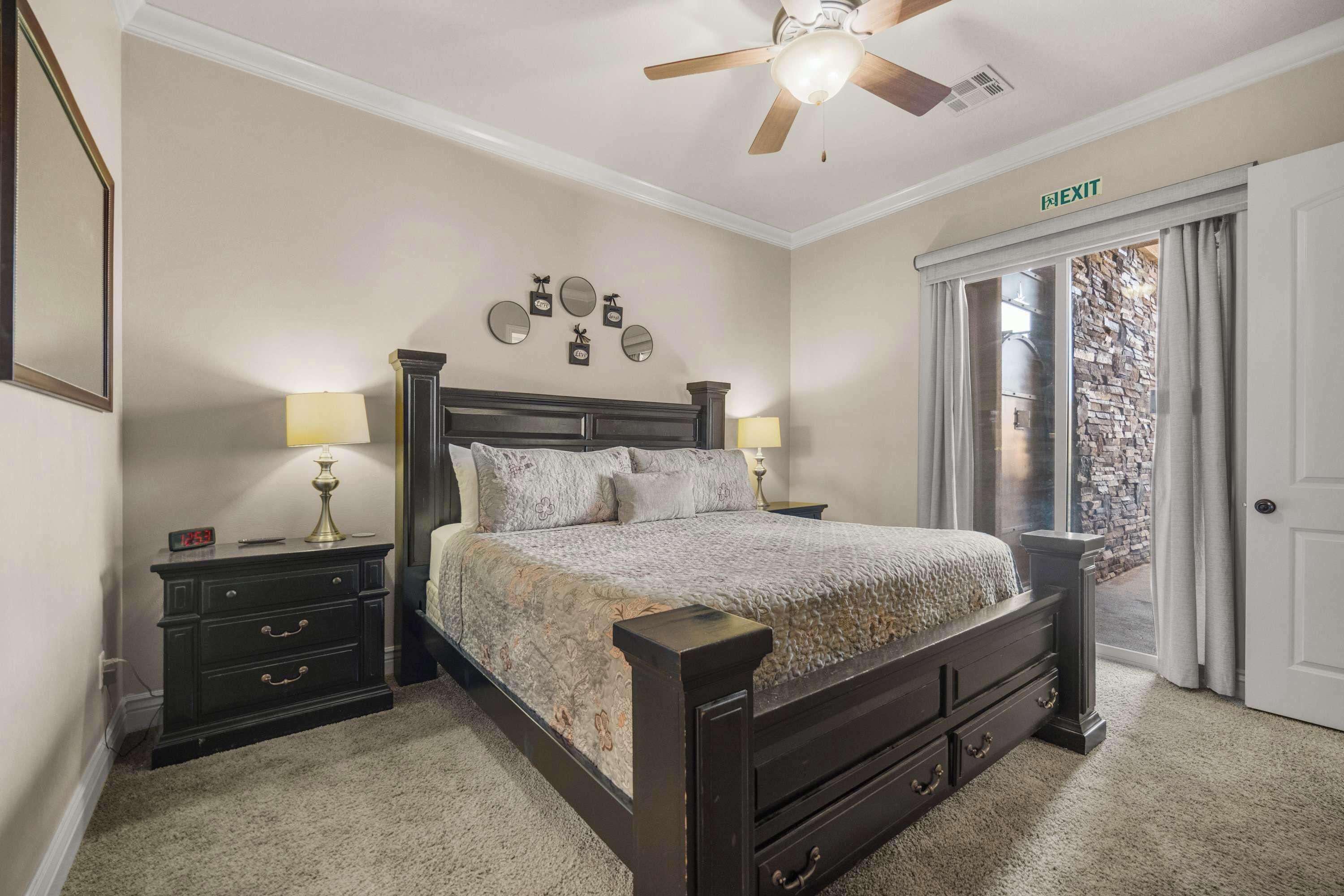 A bedroom with a bed, nightstands, lamps, a ceiling fan, and an exit sign.