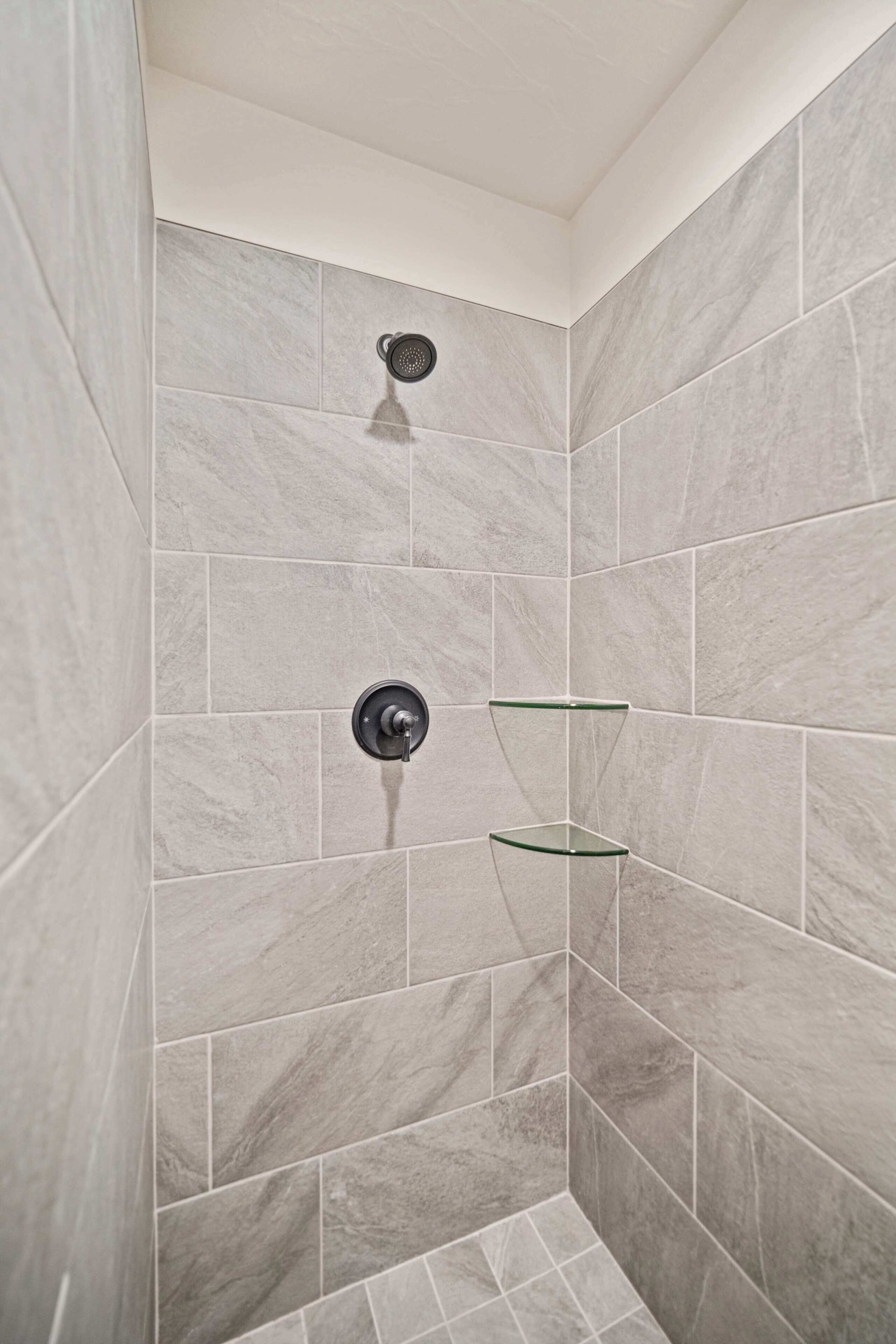 A shower with grey tiles, a showerhead, and two glass shelves.