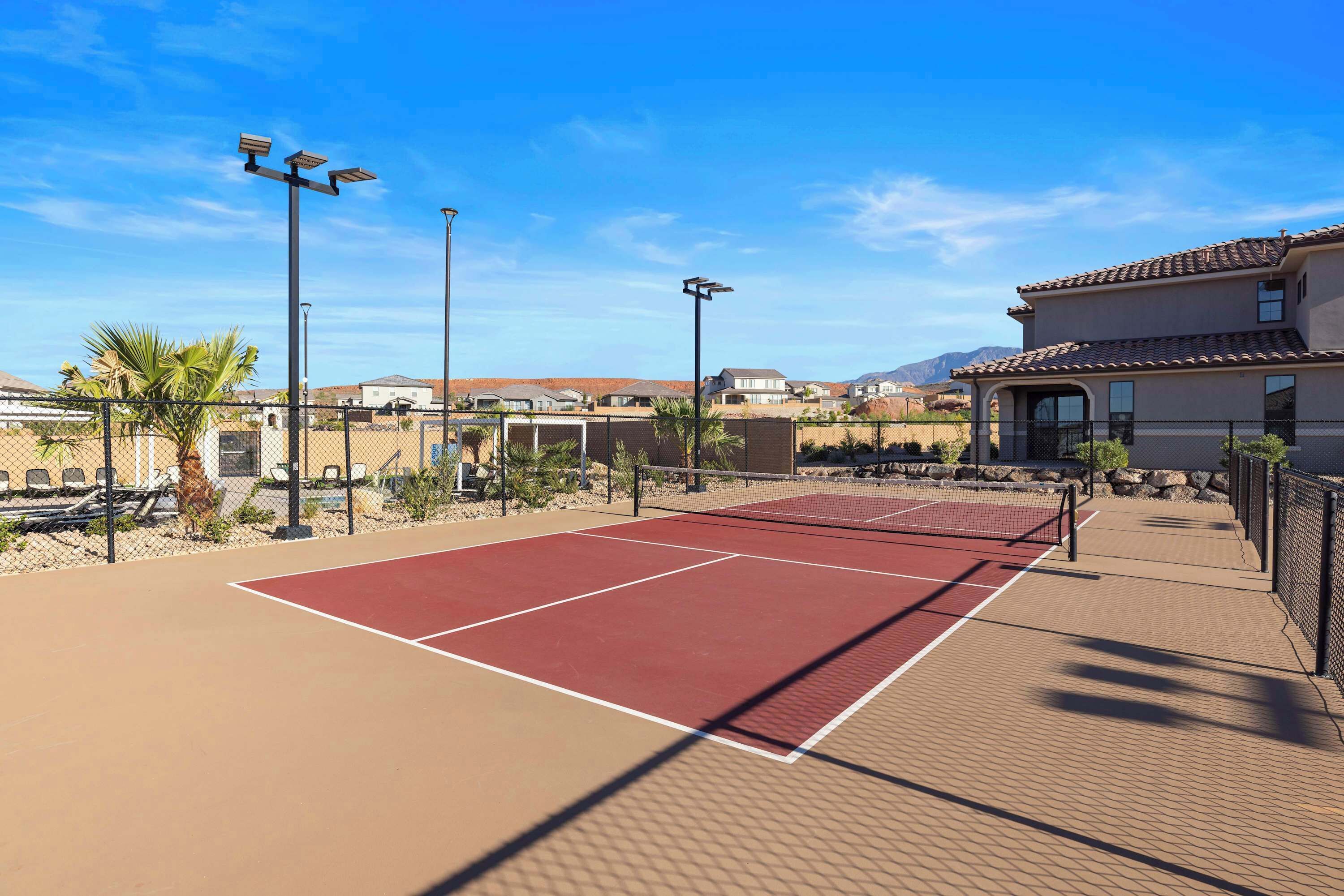 An outdoor tennis court with a red surface, surrounded by a fence, and equipped with solar-powered lights.