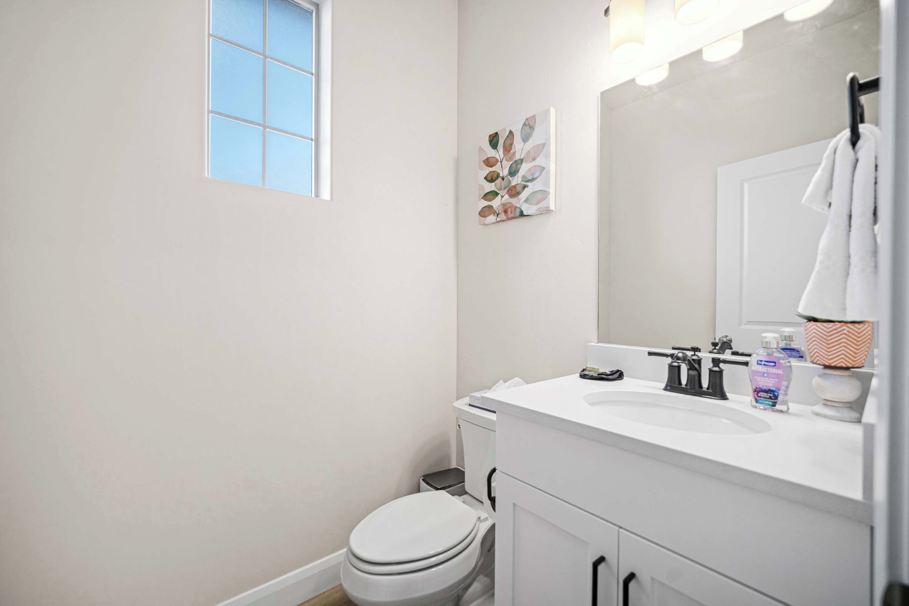 Un baño moderno con un lavamanos blanco, un espejo y un inodoro. Hay una ventana con persianas, una toalla colgada en la pared y un pequeño cubo de basura.
