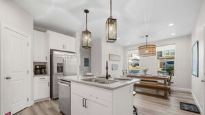 Une cuisine moderne avec des armoires blanches, des appareils en acier inoxydable et un îlot central doté d’un évier. On y trouve également des luminaires suspendus et un coin repas avec une table et des chaises en bois.