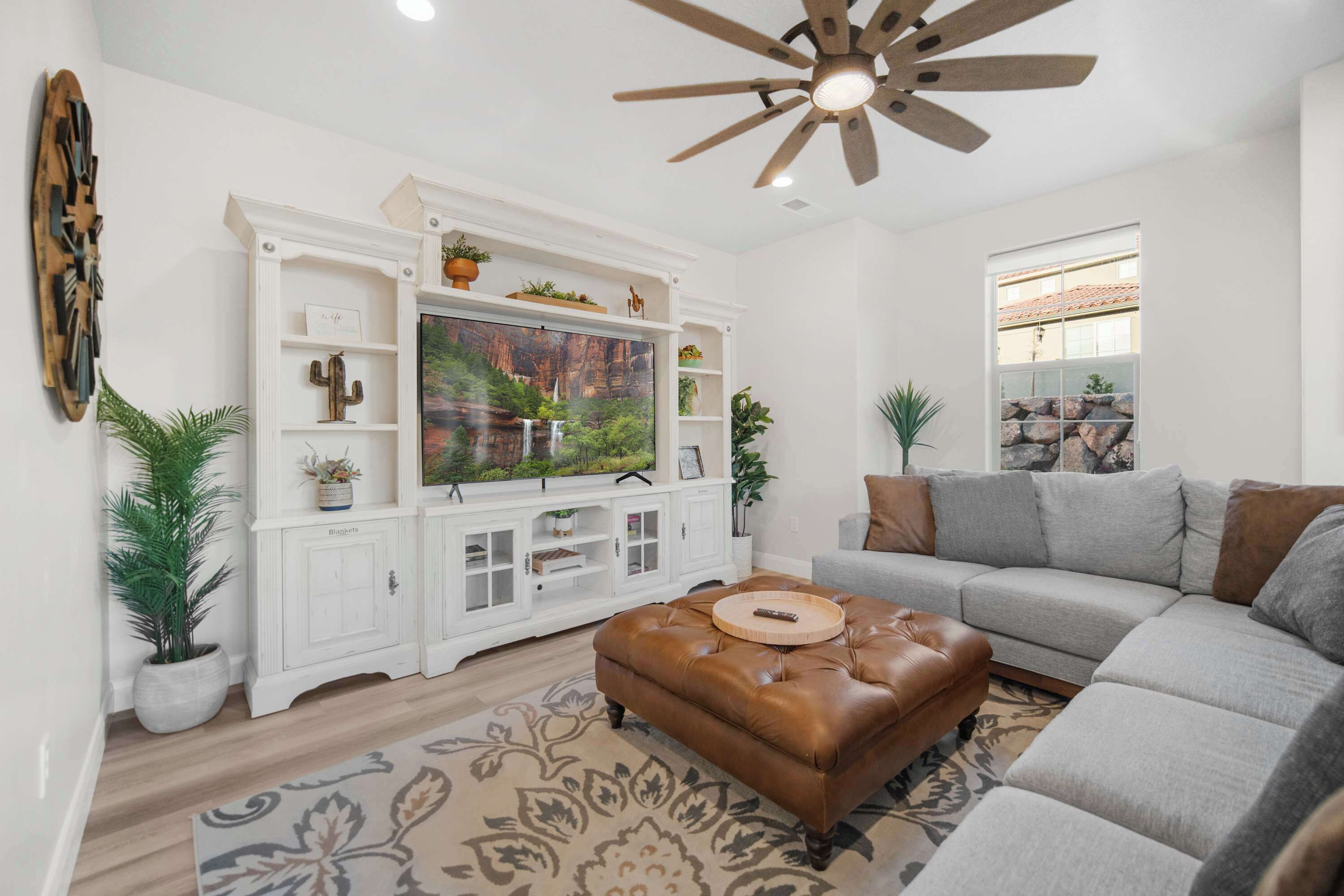 A living room with a large flat-screen TV, a brown leather ottoman, a grey sofa, and a ceiling fan.