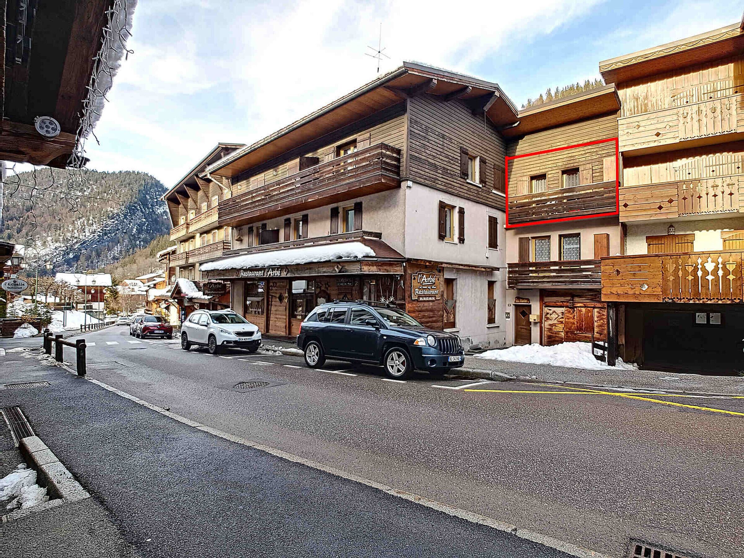 Straßenansicht mit Holzhäusern, einem Restaurant namens 'Restaurant Arbe' und geparkten Autos.