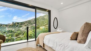 A bedroom with a large sliding glass door, a bed with white bedding, a round mirror, and a view of a hilly landscape. - Villas Pinares de San Antón, Saltwater pool, Sea, City, Mountain Views (Málaga)