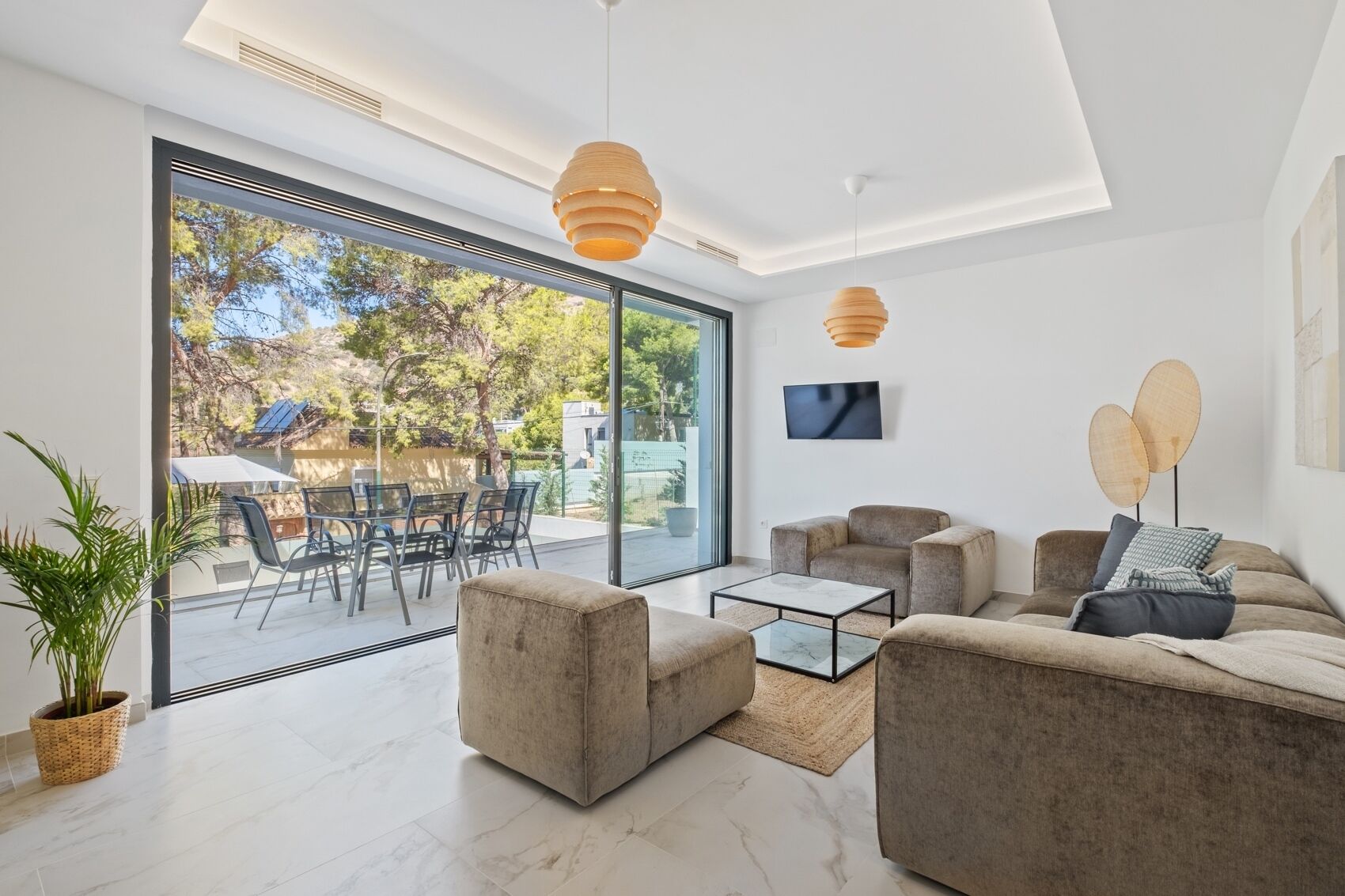 Een moderne woonkamer met een grote schuifpui die uitkomt op een terras, een zithoek, een bijzettafeltje en hanglampen.