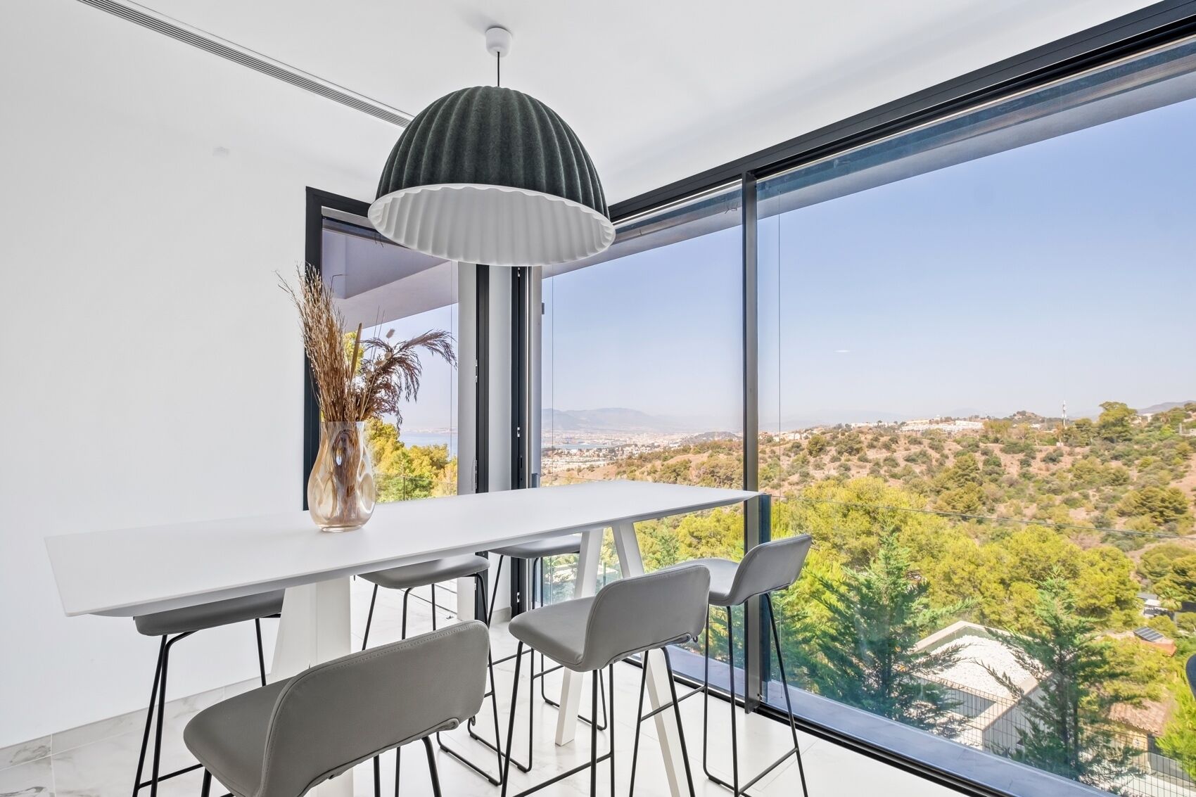 A modern dining area with a white table and grey chairs, a pendant light, and a view of a cityscape through large windows.