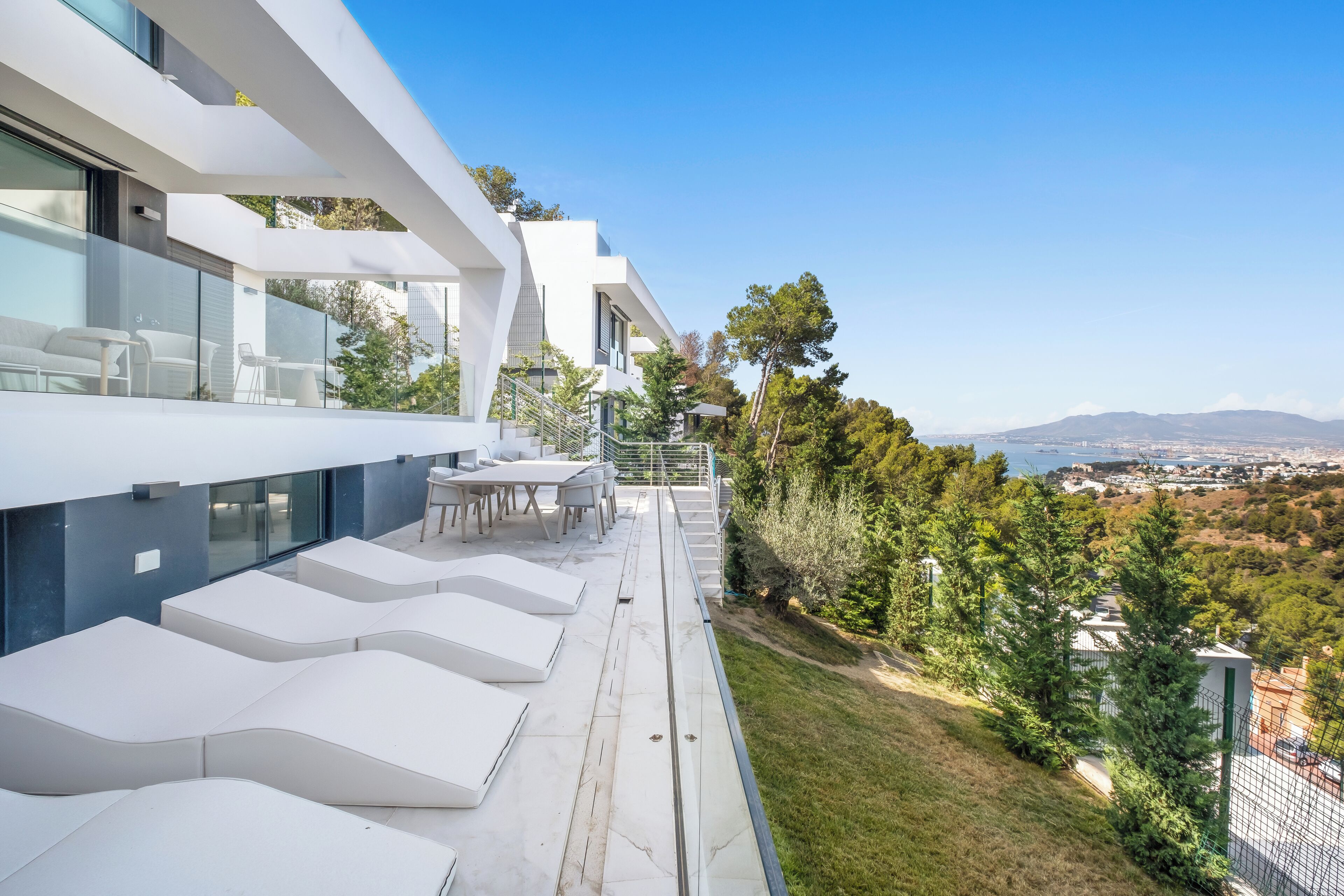 A modern outdoor terrace with white lounge chairs and a dining area, overlooking a scenic landscape with trees and a distant view of the city.