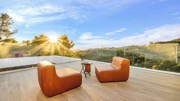 Un área de estar moderna al aire libre con dos sillones acolchados y una pequeña mesa, con vistas a un paisaje pintoresco con colinas y árboles.