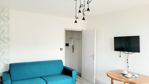 A modern living room with a teal sofa, a wooden dining table with cups and a bowl, a mounted TV, and a shower room in the background. - Renovated seafront studio in Canet, near amenities, ideal for 3 people (Canet-en-Roussillon)